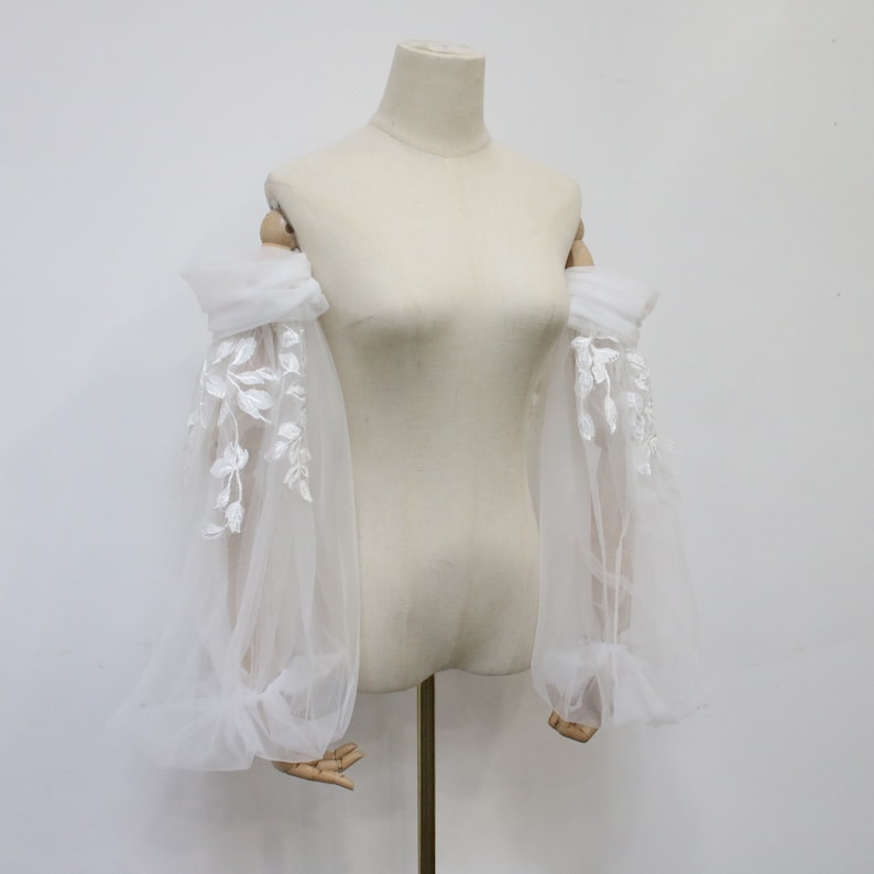 May include: White tulle bridal sleeves with delicate floral lace embroidery. The sleeves are gathered at the elbow and have a puffed look.