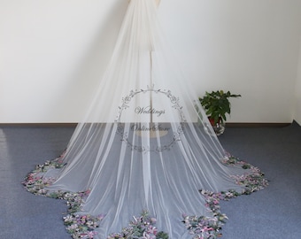 May include: A long white tulle wedding veil with a floral embroidered border in shades of purple, green, and pink. The veil is displayed on a mannequin in a white room.