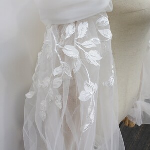 May include: White tulle fabric with delicate white floral embroidery. The fabric is sheer and has a soft, flowing texture.