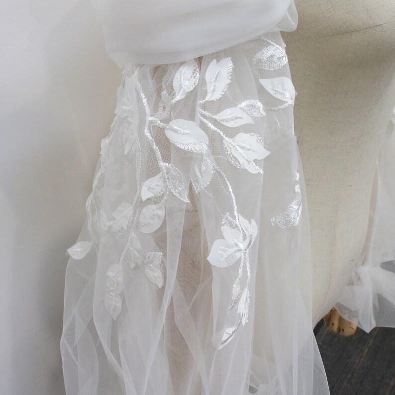May include: White tulle fabric with delicate white floral embroidery. The fabric is sheer and has a soft, flowing texture.