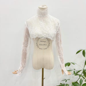Puede incluir: Un bolero de encaje blanco con mangas largas y cuello alto. El delicado diseño de encaje floral cubre todo el bolero. El bolero se exhibe en un maniquí con el texto "Weddings Online Store".