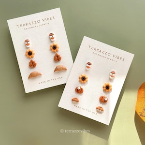 Puede incluir: Dos juegos de pendientes en tarjetas blancas con el texto "TERRAZZO VIBES handmade jewelry MADE IN THE USA". Cada juego incluye tazas de café, girasoles, hojas y cruasanes. Un juego también tiene pendientes de calabaza y tarta. El otro juego tiene pendientes de calabaza.