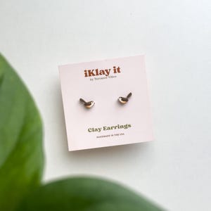 May include: A pair of small brown and white bird-shaped clay stud earrings attached to a light pink square display card. The card features the text iKlay it by Terrazzo Vibes, Clay Earrings, and HANDMADE IN THE USA printed in brown and green.