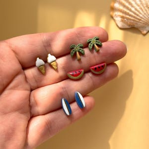 Puede incluir: Una colección de pendientes de botón con temática veraniega. Los pendientes incluyen conos de helado, palmeras, rodajas de sandía y tablas de surf. Los pendientes son de varios colores, incluyendo blanco, marrón, verde, rojo y azul. Los pendientes son pequeños y delicados.