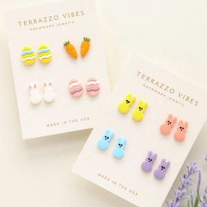 May include: Three cards with the text "TERRAZZO VIBES handmade jewelry" and "MADE IN THE USA". Each card features a set of two pastel-colored clay earrings. The first card shows a pair of carrot earrings, a pair of bunny earrings, and a pair of striped egg earrings. The second card shows a pair of pink and white striped egg earrings, a pair of yellow bunny earrings, and a pair of peach bunny earrings. The third card shows a pair of blue bunny earrings, a pair of purple bunny earrings, and a pair of yellow bunny earrings.