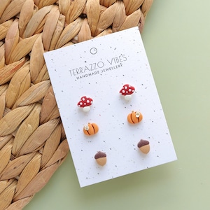 May include: A white card with the text "TERRAZZO VIBES HANDMADE JEWELLERY" displays six small earrings. The earrings are shaped like red mushrooms with white spots, orange pumpkins, and brown acorns. The earrings are on a woven background.
