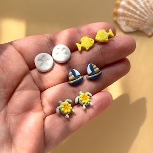 May include: A selection of miniature earrings with seaside designs. The earrings include yellow fish, blue sailboats, white sand dollars, and green turtles with yellow flowers. These earrings are ideal for anyone who loves the sea and its creatures.