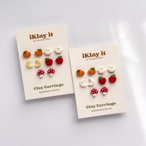 May include: Two white display cards labelled iKlay it by Terrazzo Vibes Clay Earrings Handmade in the USA, each featuring five pairs of miniature clay stud earrings shaped like orange pumpkins, white ghosts, yellow crescent moons, red apples, and red mushrooms.