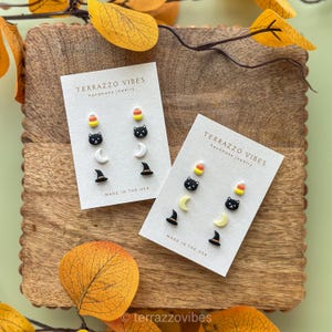 Puede incluir: Dos juegos de pendientes de Halloween en tarjetas blancas. Cada juego incluye un caramelo de maíz, una cara de gato negro, una luna creciente y un sombrero de bruja. Las tarjetas dicen "TERRAZZO VIBES handmade jewelry MADE IN THE USA."