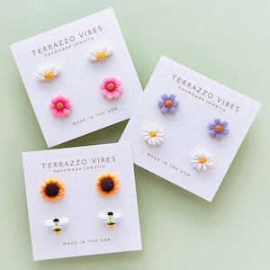 May include: Four cards with different sets of handmade polymer clay earrings. The first card features pink and white flower earrings. The second card features purple and white flower earrings. The third card features sunflower and bee earrings. Each card has the text "TERRAZZO VIBES handmade jewelry" and "MADE IN THE USA".