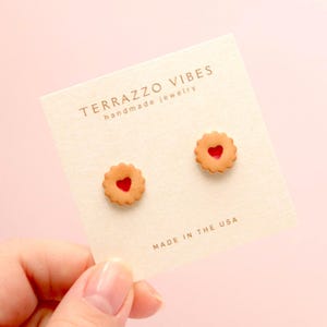 May include: A pair of small, red and brown cookie earrings with a heart-shaped jelly filling. The earrings are on a white card with the text "TERRAZZO VIBES handmade jewelry" and "MADE IN THE USA".