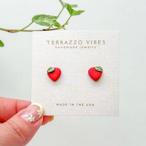 Puede incluir: Un par de pendientes de botón de fresa roja y verde en una tarjeta blanca con el texto "TERRAZZO VIBES handmade jewelry" y "MADE IN THE USA".