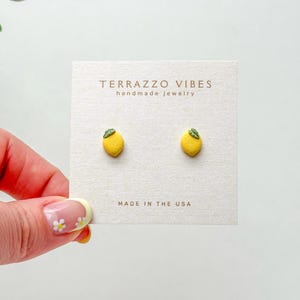 Puede incluir: Un par de pendientes de botón de limón amarillo con hojas verdes. Los pendientes están hechos a mano y dicen "Terrazzo Vibes handmade jewelry" y "Made in the USA" en la tarjeta.
