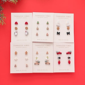May include: Six sets of handmade holiday-themed earrings on white cards. Designs include a red hat, Christmas tree, milk carton, cookie, bow, gingerbread man, snowflake, snowman, car with tree, and Christmas light. The cards read "TERRAZZO VIBES handmade jewelry MADE IN THE USA."