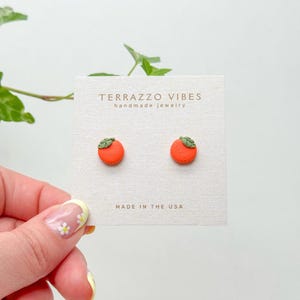 Puede incluir: Un par de pendientes de botón de fruta naranja con hojas verdes. Los pendientes están en una tarjeta blanca con el texto "Terrazzo Vibes handmade jewelry" y "Made in the USA".