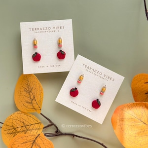 May include: Two pairs of handmade stud earrings on white cards. One pair features a yellow pencil with a pink eraser, and a red apple. The other pair has the same design. The cards read "TERRAZZO VIBES handmade jewelry MADE IN THE USA."