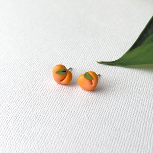 Puede incluir: Un par de pendientes de botón con forma de melocotón naranja con pequeños detalles de hojas verdes. Los pendientes están sobre un fondo blanco, con una hoja verde en la esquina superior derecha. Un accesorio divertido y único.