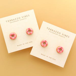 Puede incluir: Dos pares de pendientes de botón de donut en tarjetas blancas. Las tarjetas tienen el texto "TERRAZZO VIBES handmade jewelry" y "MADE IN THE USA". Los donuts son rosados con glaseado rosado y chispas marrones.