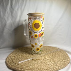 May include: A clear glass tumbler with a bamboo lid and a straw. The tumbler features a sunflower and butterfly design with the words "sunshine", "kindness", "beauty", "happiness", "joy", and "love you".