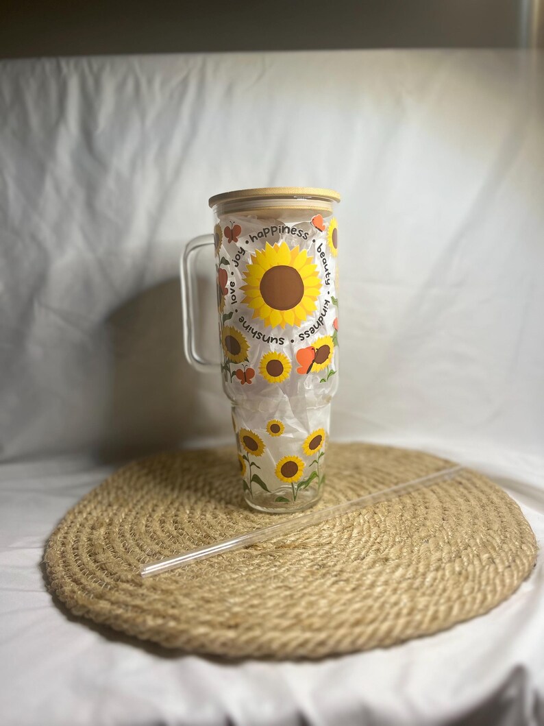 May include: A clear glass tumbler with a bamboo lid and a straw. The tumbler features a sunflower and butterfly design with the words "sunshine", "kindness", "beauty", "happiness", "joy", and "love you".