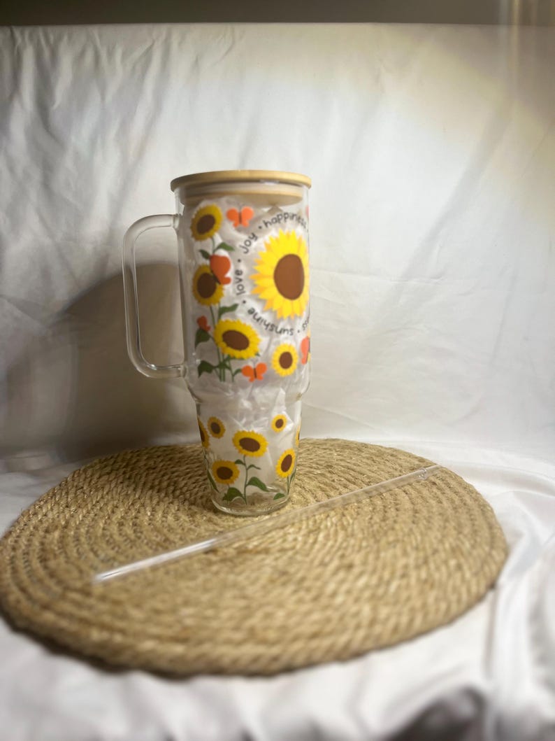 May include: A clear glass tumbler with a bamboo lid and a straw. The tumbler features a sunflower and butterfly design with the text "love, joy, happiness, sunshine".