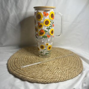 May include: A clear glass tumbler with a wooden lid and a handle. The tumbler is decorated with a sunflower and butterfly design. The tumbler is sitting on a woven straw mat.