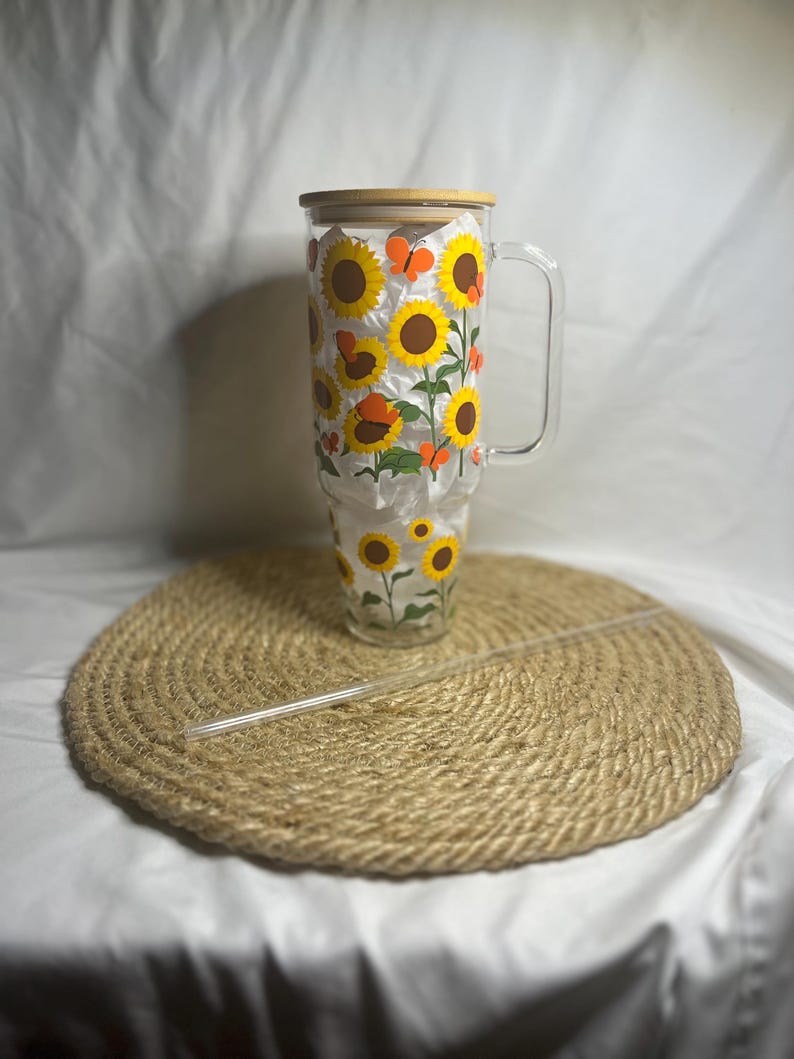 May include: A clear glass tumbler with a wooden lid and a handle. The tumbler is decorated with a sunflower and butterfly design. The tumbler is sitting on a woven straw mat.