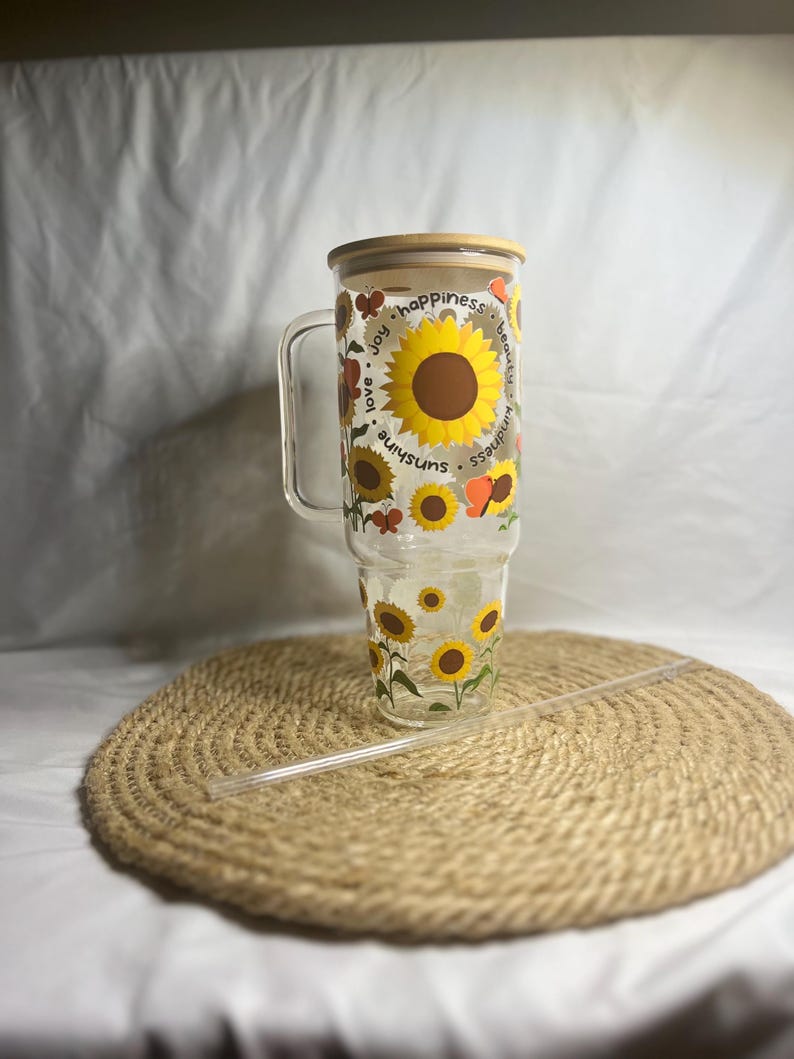 May include: A clear glass tumbler with a wooden lid and a straw. The tumbler features a sunflower design with the words "sunshine, love, joy, happiness, beauty, kindness" printed around the sunflowers.