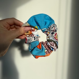 Peut inclure: Un chouchou coloré avec un mélange de motifs et de couleurs, notamment du bleu, de l'orange et des motifs floraux. Le tissu est froncé et élastiqué, créant une forme circulaire. Cet accessoire pour cheveux est parfait.