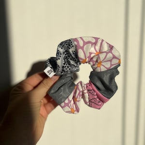 Può includere: Un colorato elastico per capelli con un mix di fantasie e colori. L'elastico presenta sezioni di tessuto paisley bianco e nero, grigio, rosa e con stampa a margherite bianche. È attaccata una piccola etichetta.