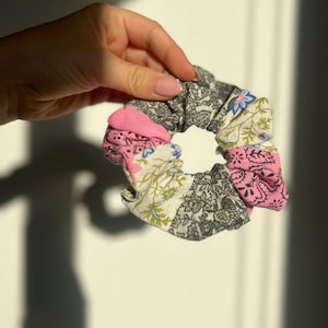 Peut inclure: Un chouchou coloré avec un motif patchwork. Le tissu présente des motifs floraux roses, blancs, gris et bleus. Le chouchou est tenu dans une main, mettant en valeur ses couleurs et textures vives.