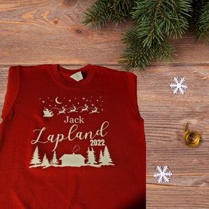 Pu&ograve; includere: Maglione rosso di Natale con un design bianco di una capanna, alberi e renne che volano con Babbo Natale. Il testo "Jack Lapland 2022" &egrave; sul maglione.