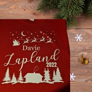Pu&ograve; includere: Maglione rosso di Natale con testo bianco che dice "Davie Lapland 2022". Il maglione presenta una silhouette bianca di Babbo Natale in una slitta trainata da renne, una luna crescente e stelle.