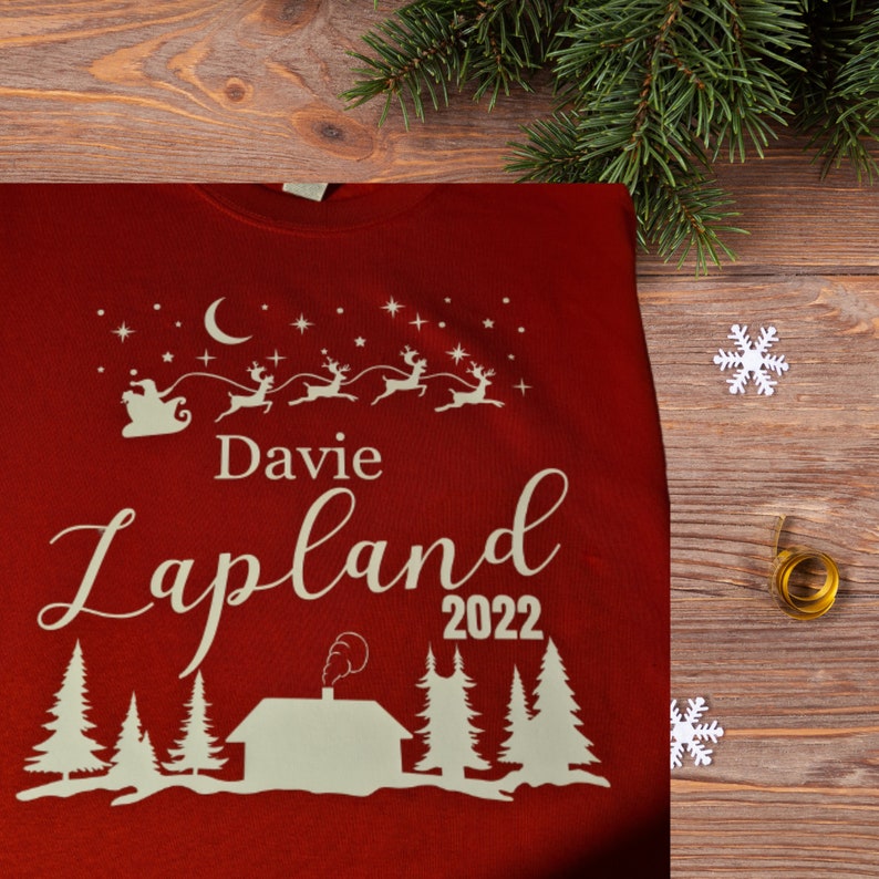 Pu&ograve; includere: Maglione rosso di Natale con testo bianco che dice "Davie Lapland 2022". Il maglione presenta una silhouette bianca di Babbo Natale in una slitta trainata da renne, una luna crescente e stelle.