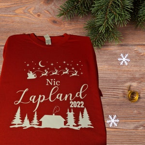 Pu&ograve; includere: Maglietta a maniche lunghe rossa con un design bianco di Babbo Natale su una slitta trainata da renne, una luna crescente, stelle, una capanna con camino, alberi e il testo "Nic Lapland 2022".