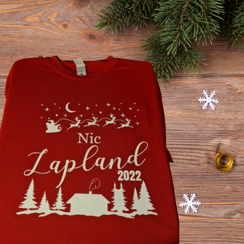 Pu&ograve; includere: Maglietta a maniche lunghe rossa con un design bianco di Babbo Natale su una slitta trainata da renne, una luna crescente, stelle, una capanna con camino, alberi e il testo "Nic Lapland 2022".
