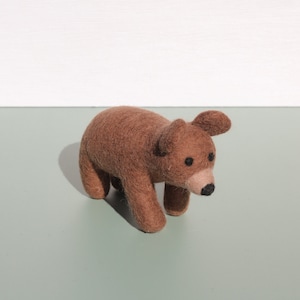 May include: A brown felt bear toy with black eyes and a brown nose. The bear is standing on a light green surface.