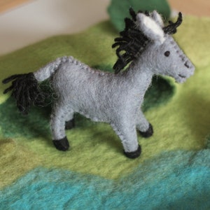 May include: A handmade gray felt donkey figurine with black mane, tail, and hooves. The donkey is standing on a colorful felt landscape of green, yellow, and blue. The donkey is a charming, handcrafted item.