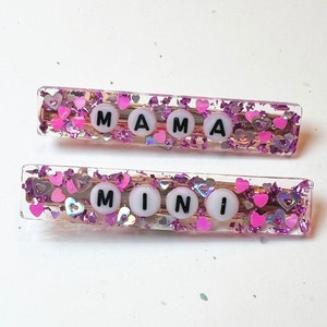 May include: Two pink hair clips with clear resin and glitter. The top clip says "MAMA" and the bottom clip says "MINI".