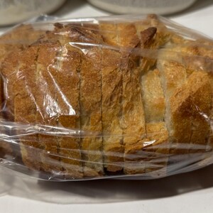 May include: A loaf of sliced bread, encased in a clear plastic bag. The bread has a golden-brown crust and a light interior, with visible slices. The packaging is sealed with a twist tie. The bread appears fresh and ready to eat.