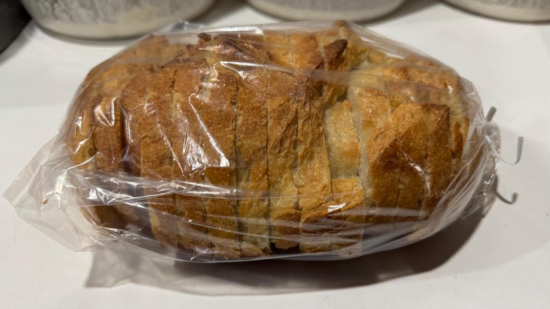 May include: A loaf of sliced bread, encased in a clear plastic bag. The bread has a golden-brown crust and a light interior, with visible slices. The packaging is sealed with a twist tie. The bread appears fresh and ready to eat.