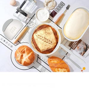 May include: Overhead shot of sourdough bread baking supplies. Includes a loaf of bread with a leaf design, a bowl of flour, a dough scraper, a whisk, a measuring jar, and a loaf of dough in a container.
