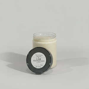 May include: A clear glass jar filled with a creamy, off-white substance, likely a sourdough starter. The black lid, with white text reading "Live Sourdough Starter," is placed next to the jar. The jar and lid are on a light background.