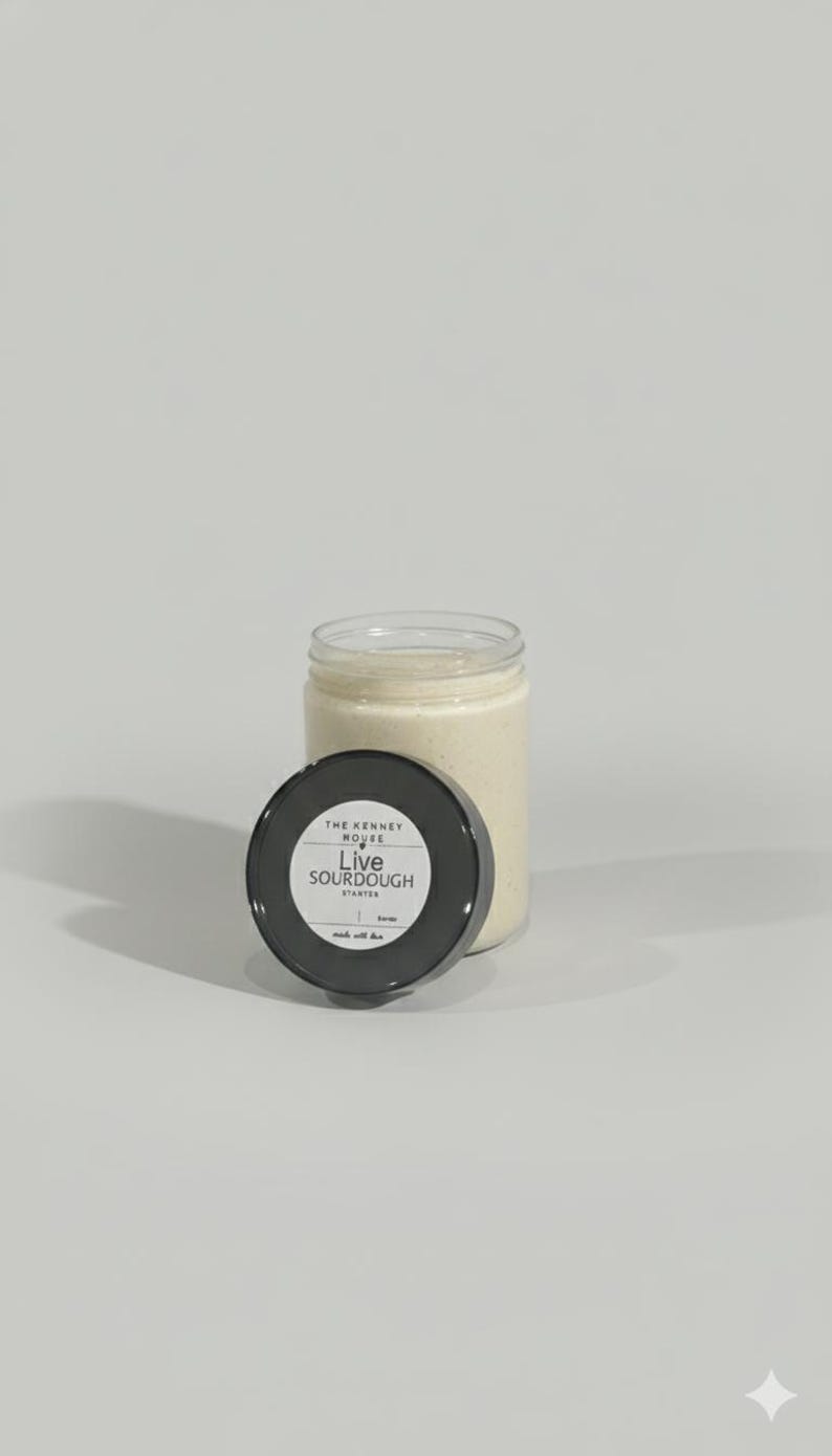 May include: A clear glass jar filled with a creamy, off-white substance, likely a sourdough starter. The black lid, with white text reading "Live Sourdough Starter," is placed next to the jar. The jar and lid are on a light background.