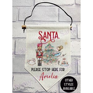 May include: A festive Christmas banner with the word "SANTA" in red script. The banner features a nutcracker, a ballerina, and a Christmas tree. The text "PLEASE STOP HERE FOR Amelia" is below the illustration. A black cord is attached for hanging.