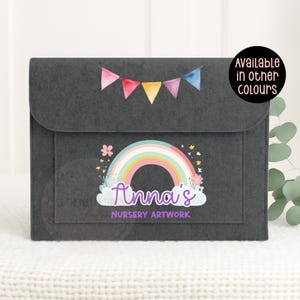 May include: A dark grey felt portfolio with a flap closure. It features a colourful rainbow design with the name "Anna's" and the text "Nursery Artwork." A banner of small flags is above the rainbow. The text "Available in other colours" is in a circle.