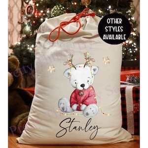 May include: A beige Christmas sack with a drawstring and red cord. It features a watercolour illustration of a white bear wearing a red sweater and antlers with Christmas lights. The name "Stanley" is written in black script.