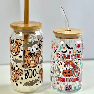 May include: Two glass Halloween themed tumblers with lids and straws. The tumbler on the left has a Mickey Mouse design with pumpkins and bats. The tumbler on the right has a rainbow, skull, and pumpkin design with the text "Here for the boos". The tumbler on the left has a bamboo straw and the tumbler on the right has a glass straw.