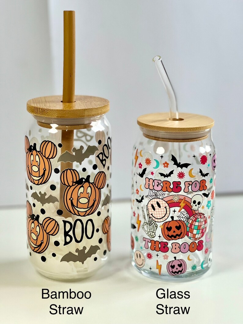 May include: Two glass Halloween themed tumblers with lids and straws. The tumbler on the left has a Mickey Mouse design with pumpkins and bats. The tumbler on the right has a rainbow, skull, and pumpkin design with the text "Here for the boos". The tumbler on the left has a bamboo straw and the tumbler on the right has a glass straw.