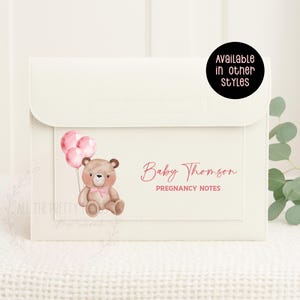 May include: A cream-coloured pregnancy notes organiser with a flap closure. It features a watercolour illustration of a teddy bear holding pink balloons. The text "Baby Thomson" and "Pregnancy Notes" are in pink script.